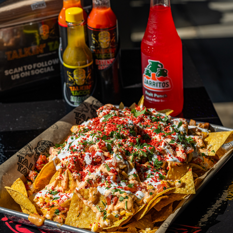 Talkin' Tacos nachos with red seasoning, cilantro, and sauces, alongside hot sauces and a Jarritos soda.