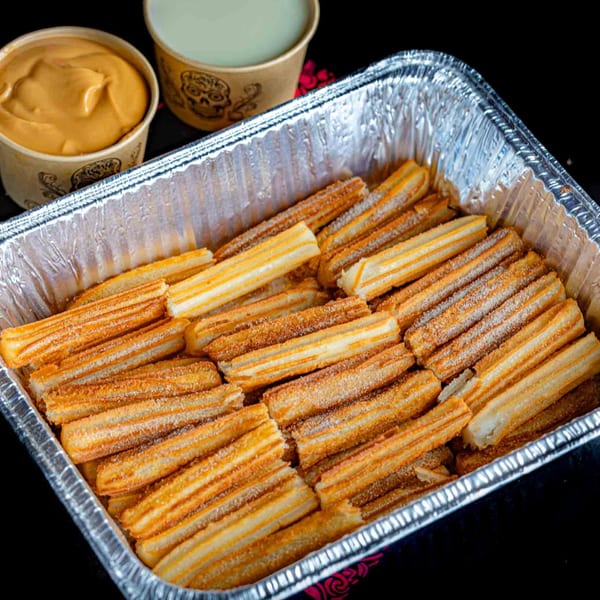 A foil tray filled with golden-brown churros dusted with cinnamon sugar, alongside two cups of dipping sauces.