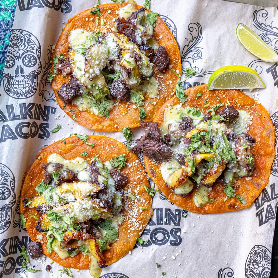 Three Talkin' Tacos birria tacos with meat, cheese, and cilantro, on branded paper with lime wedges.