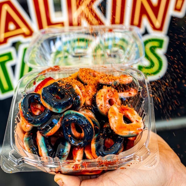A hand holds a clear plastic container filled with mini donuts, some blue, some orange, covered in chili powder.