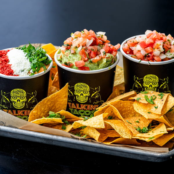 Three Talkin' Tacos cups filled with various toppings and dips, surrounded by tortilla chips on a tray.