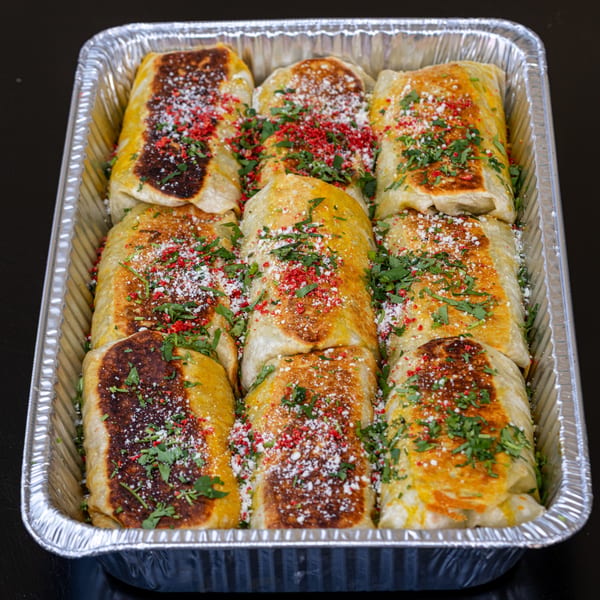 Nine golden-brown burritos, topped with red spice, white cheese, and green herbs, in a foil tray.