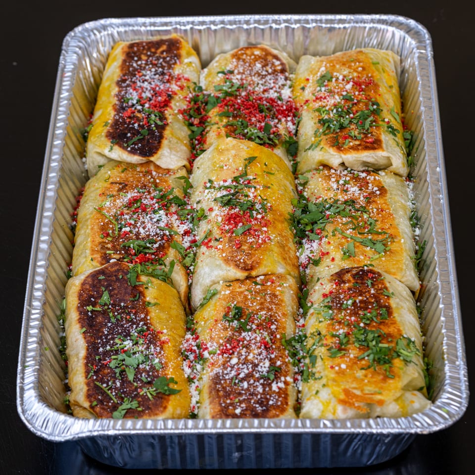 Nine golden-brown burritos, topped with red spice, white cheese, and green herbs, in a foil tray.
