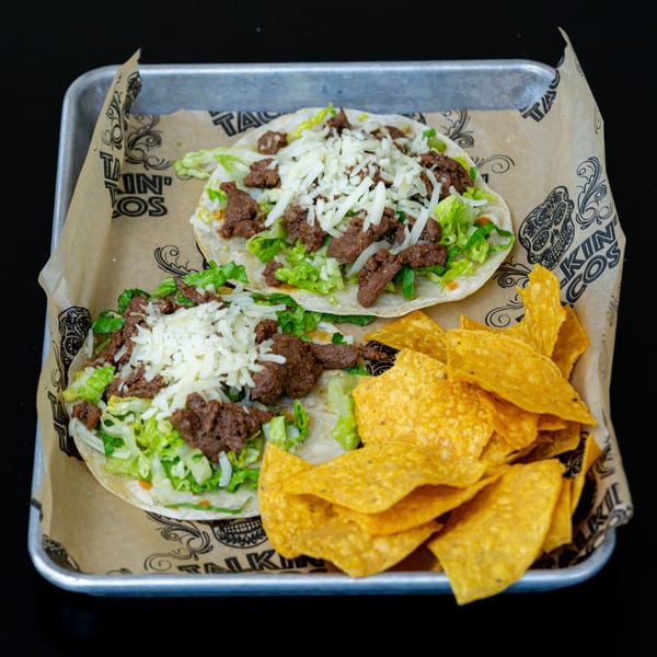 Two Talkin' Tacos with beef, lettuce, and cheese, served with tortilla chips on a metal tray.