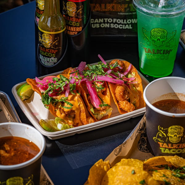 Three Talkin' Tacos with sauce, pickled onions, and cilantro, alongside hot sauce and a green drink.