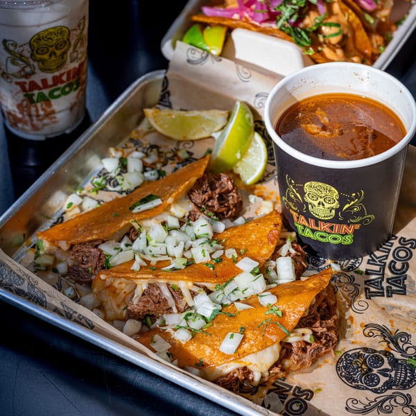 Three Talkin' Tacos birria tacos with onions and cilantro, a cup of consommé, and lime wedges.