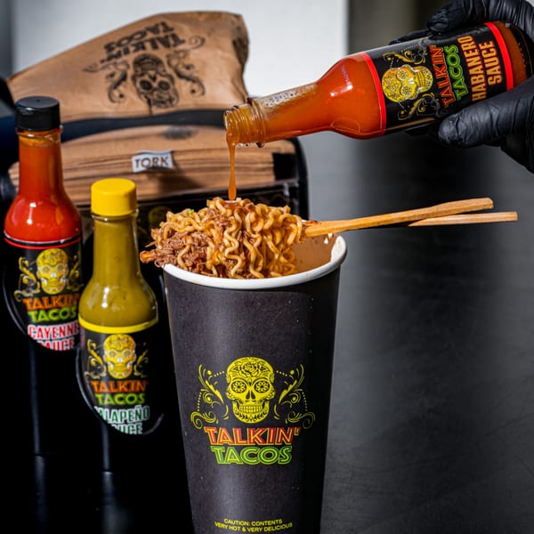 Hand in black glove pouring Talkin' Tacos Habanero sauce onto noodles in a branded cup, with other sauces nearby.