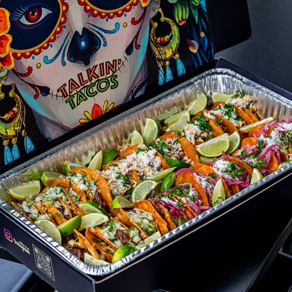 A catering tray of Talkin' Tacos birria tacos with lime wedges, cilantro, and onions.