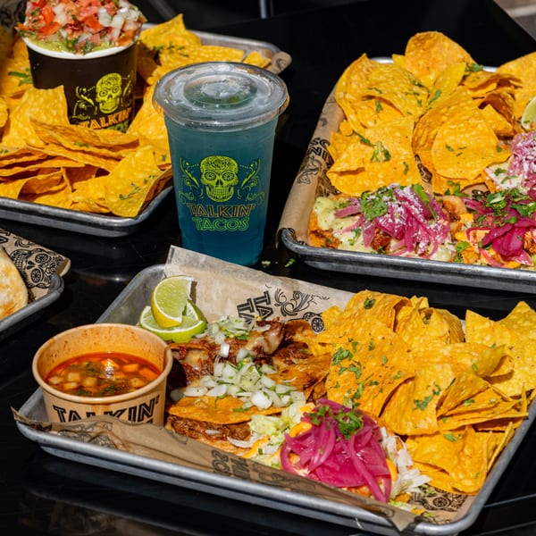 Talkin' Tacos spread with birria tacos, nachos, guacamole, and a blue drink on metal trays.