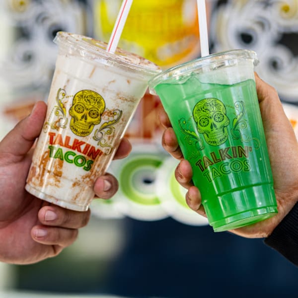 Two hands holding up Talkin' Tacos branded cups, one with a horchata drink and one with a green drink.