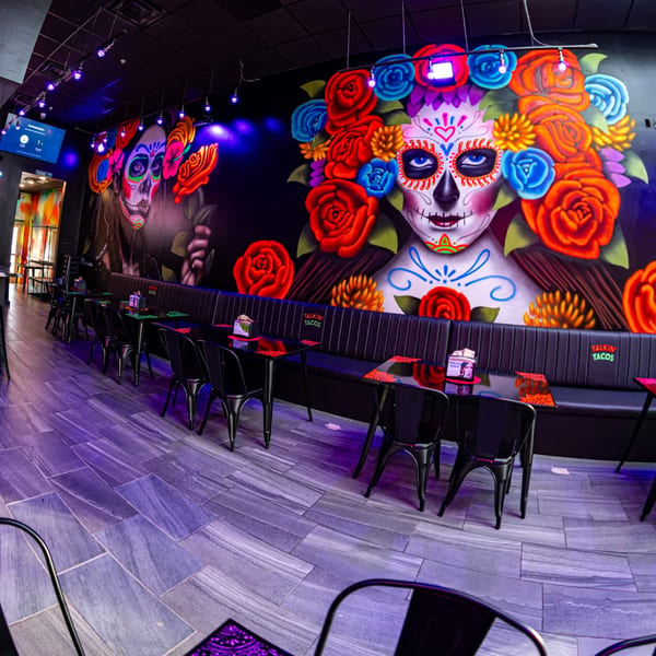 Talkin' Tacos restaurant interior with vibrant Day of the Dead murals, black tables, and purple lighting.