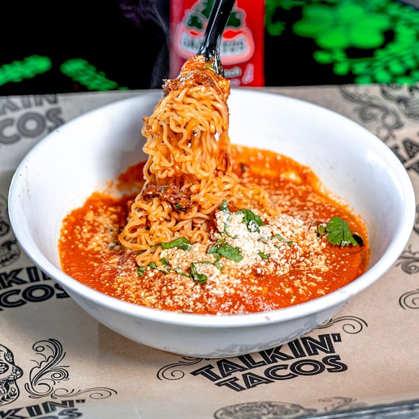 A fork lifts ramen noodles from a white bowl of red sauce, cheese, and cilantro.
