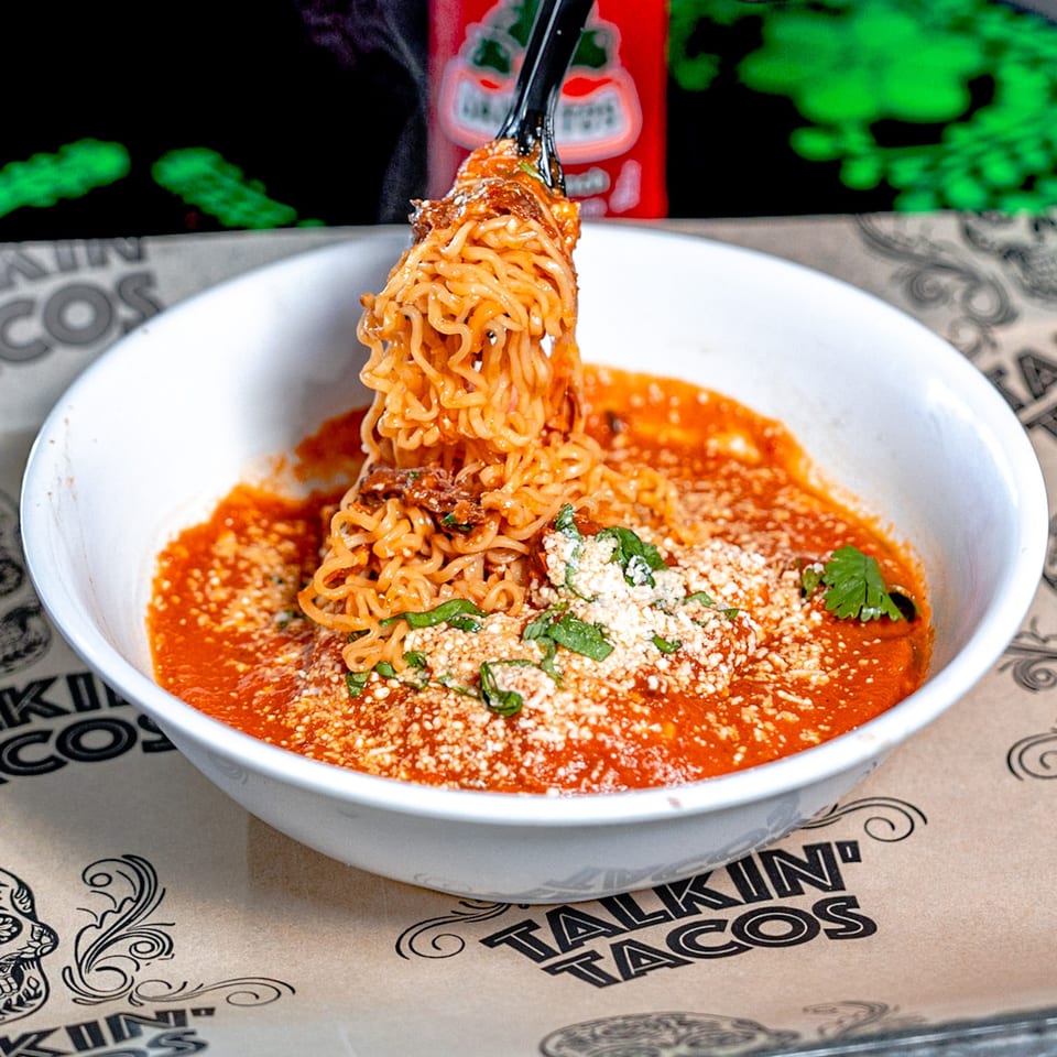 Tongs lifting ramen noodles from a white bowl of red sauce, cheese, and cilantro.