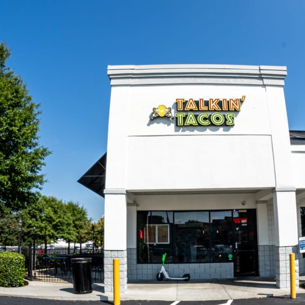 Exterior of Talkin' Tacos restaurant with outdoor seating, a scooter, and blue sky.