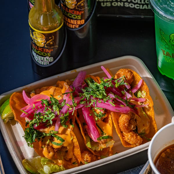 Three Talkin' Tacos with shrimp, sauce, pickled onions, and cilantro, alongside hot sauce and a drink.