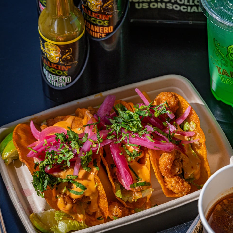 Three Talkin' Tacos with shrimp, sauce, pickled onions, and cilantro, alongside hot sauce and a drink.