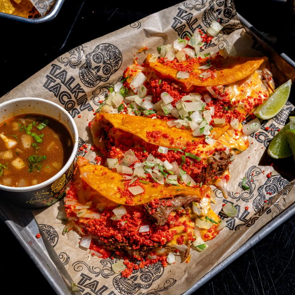 Four Talkin' Tacos birria tacos with cheese, onions, and red seasoning, served with consommé and lime.