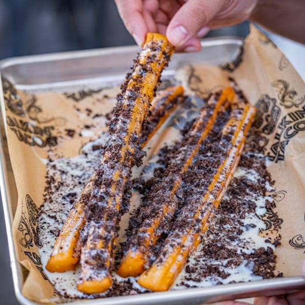 Hand picking up a churro covered in white icing and dark cookie crumbles from a tray.