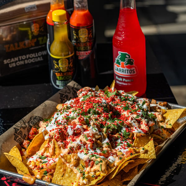 Talkin' Tacos loaded nachos with sauces, cilantro, and red sprinkles, alongside hot sauce bottles and a Jarritos.