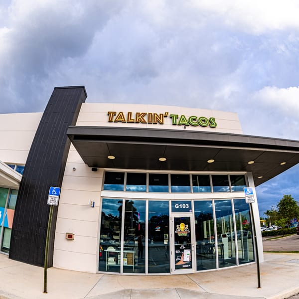 Exterior of Talkin' Tacos restaurant with a large sign, glass windows, and a dark, cloudy sky.
