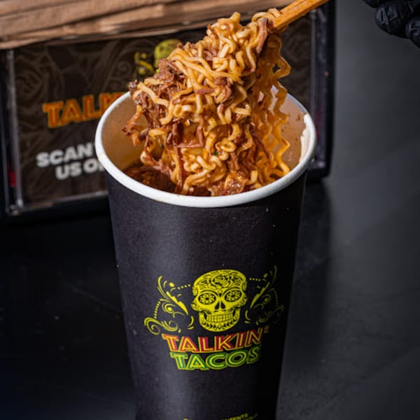 Chopsticks lifting saucy noodles and shredded beef from a black Talkin' Tacos cup.