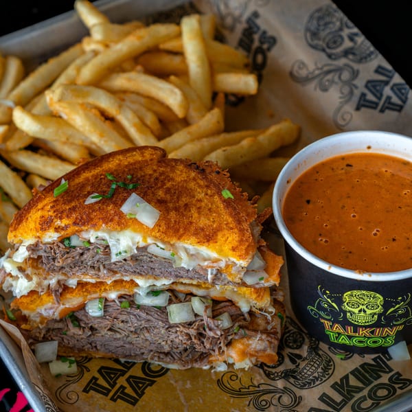 Talkin' Tacos birria grilled cheese with fries and a cup of consommé on branded paper.