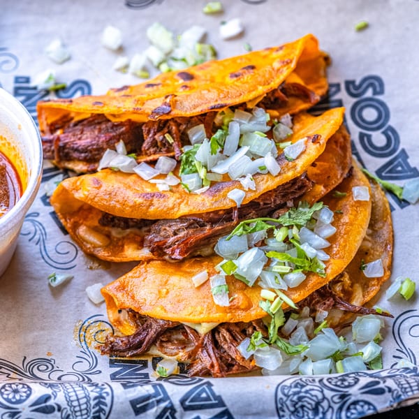 Three Talkin' Tacos birria tacos with shredded beef, cheese, onions, and cilantro, with a side of consommé.