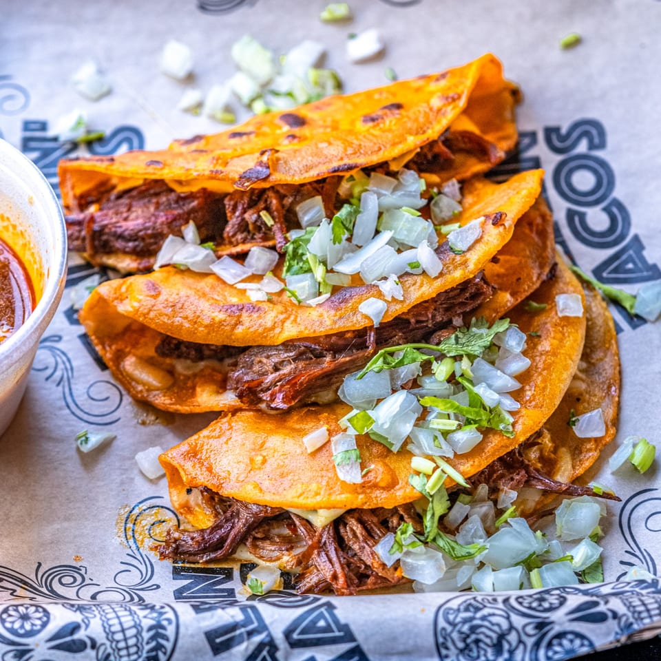 Three Talkin' Tacos birria tacos with shredded beef, cheese, onions, and cilantro, with a side of consommé.