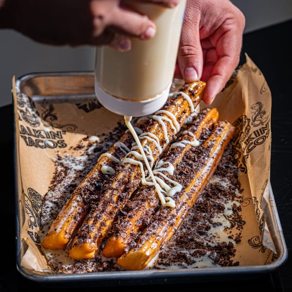 A hand squeezes a bottle, drizzling white icing over Talkin' Tacos churros with cookie crumbles.