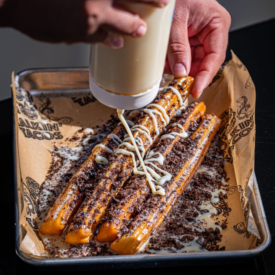A hand squeezes a bottle, drizzling white icing over Talkin' Tacos churros with cookie crumbles.