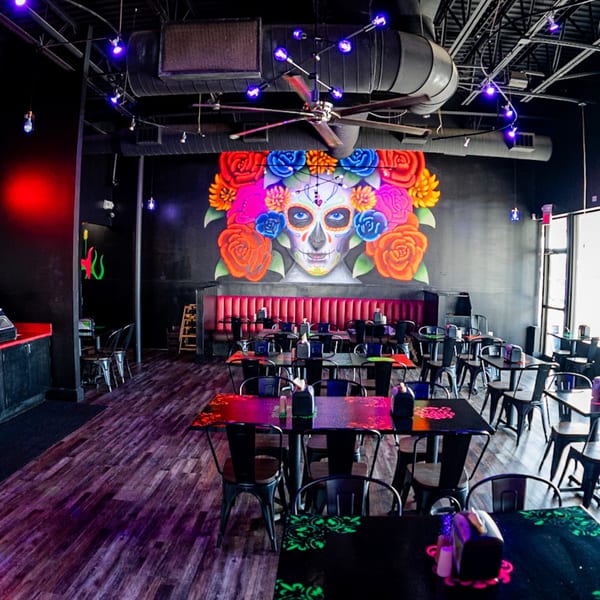 The interior of Talkin' Tacos restaurant with a large colorful sugar skull mural and dining tables.