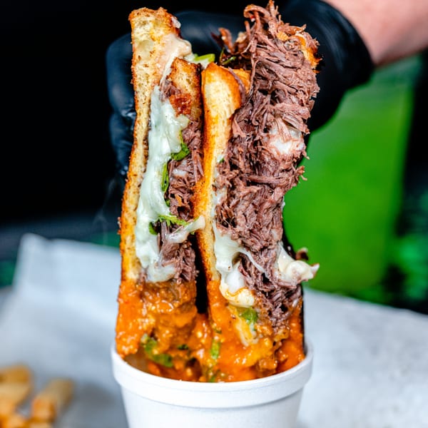 A hand in a black glove holds up a large, cheesy birria taco from Talkin' Tacos.
